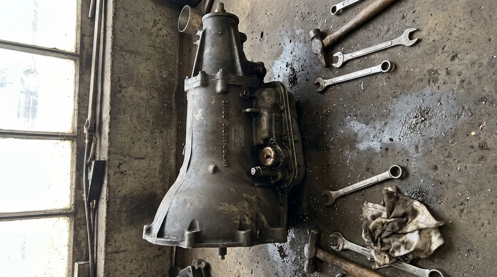 Cast iron Chevrolet Powerglide transmission on a workshop floor beside a 1950s car, bell housing facing the camera